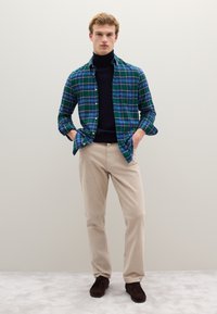 Camisa de franela a cuadros verde y azul sobre un suéter de cuello alto negro, combinado con pantalones de pana beige y zapatos marrón oscuro.