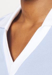 Polo shirt azzurro chiaro con colletto bianco, caratterizzato da un tessuto strutturato e un scollo a V. Vista ravvicinata dell'area superiore del petto.