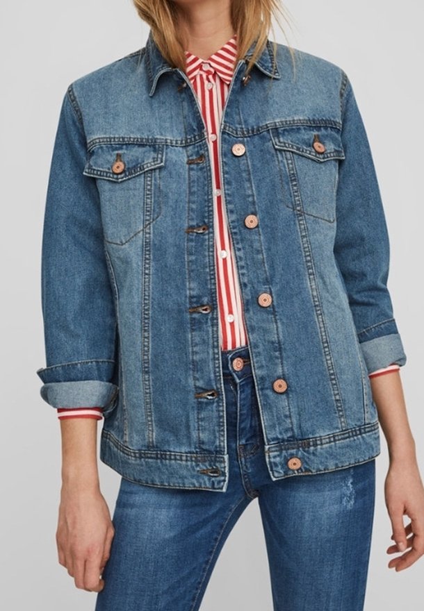 Kvinna iklädd en blå denimjacka med upprullade ärmar över en röd och vitrandig skjorta med knappar och blå jeans.