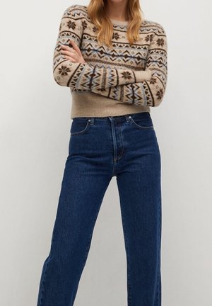 Femme les bras croisés portant un pull beige à motifs et un jean bleu foncé taille haute sur un fond uni.