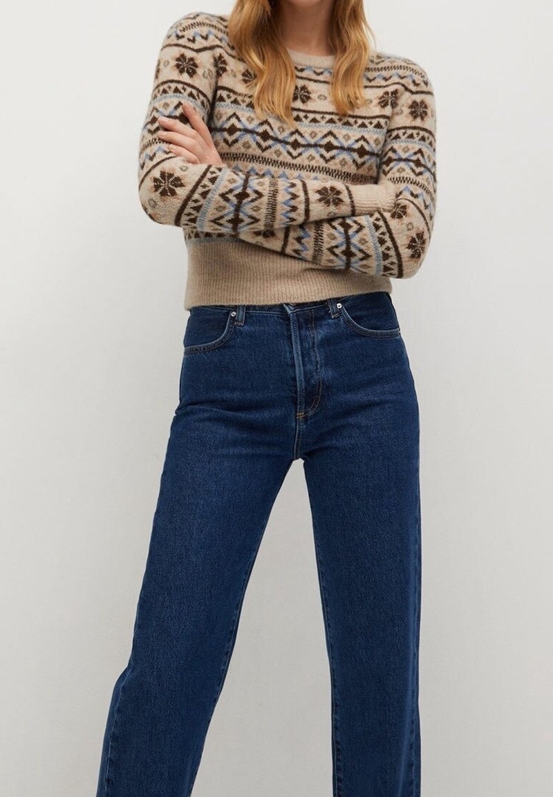 Femme les bras croisés portant un pull beige à motifs et un jean bleu foncé taille haute sur un fond uni.