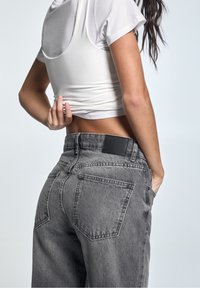 Jeans de mezclilla gris con cintura alta y cinco bolsillos, con una etiqueta de cintura negra y un corte relajado. Combinados con un top corto blanco.