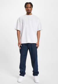 White oversized cotton t-shirt with dropped shoulders, paired with dark blue jeans and white sneakers, standing on a neutral background.