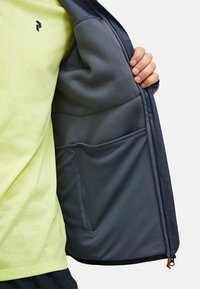 Gray jacket interior with a soft texture, featuring a large pocket. A bright yellow T-shirt with a logo is visible underneath.