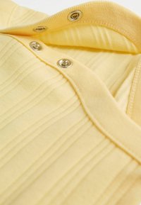 Dzeltens īsslīpains krekls ar vertikālu svītru tekstūru, ribjotu apkakli un sudraba spiedpogām. Mīksts kokvilnas materiāls ar smalku spīdumu.