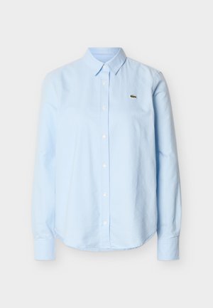 Chemise bleu clair à manches longues, boutonnée, avec col et petit logo de crocodile vert sur le côté gauche de la poitrine.