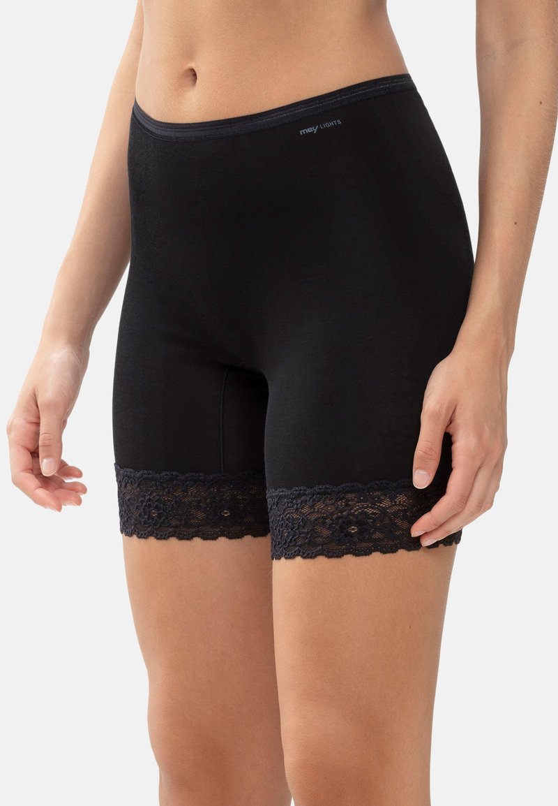 Nainen, jolla on ylhäältä avonaiset mustat korkeat shortit, joissa on kukkakuvioinen pitsireuna reisillä, seisoo yksinkertaista taustaa vasten.