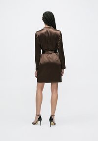 Brązowa sukienka z satyny z długimi rękawami, kołnierzykiem i pasem w talii w tym samym kolorze. Czarne buty na wysokim obcasie. Jednolite tło.