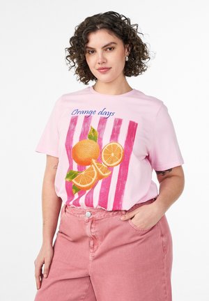 Kvinde med krøllet hår iført en pink "Orange days" t-shirt med appelsingrafik og lyserøde bukser, poserer med den ene hånd i lommen.