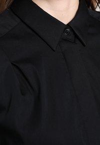 Black shirt featuring a classic collar, button closure, and a smooth texture. Includes subtle pleats near the shoulder seams.