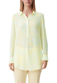 Donna che indossa una blusa a maniche lunghe di un pallido giallo con un sottile motivo di foglie azzurro chiaro e pantaloni color crema, in piedi contro uno sfondo bianco.