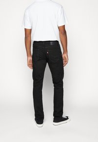 Černé džíny slim fit vyrobené z denimu, s koženou nášivkou logo, pěti kapsami a decentními švy, nošeny s bílým tričkem.
