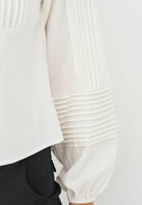 Hvid langærmet bluse med plissering på ærmerne. Glat tekstur, let løs pasform, parret med et sort plagg.