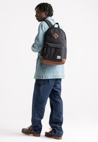 Zaino in tessuto nero con base in pelle marrone, toppe a rombo, tasca frontale con zip e spallacci regolabili. Il modello indossa una giacca di jeans e pantaloni blu navy.