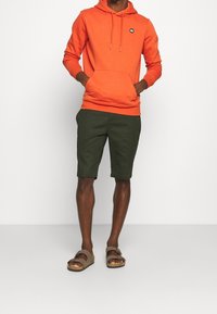 Orange hoodie med kängurufickor, ihopkopplad med mörkgröna shorts. Bruna sandaler med två justerbara remmar fulländar outfiten.
