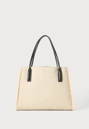 Sac à main beige avec des poignées en cuir noir, texture douce et accents en shearling sur les côtés. Présente un logo embossé sur le devant.