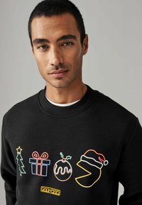 Sudadera negra con diseños coloridos bordados: árbol de Navidad, regalo, pudín, acebo y Pac-Man con un sombrero de Santa.