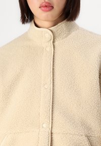 Beige fleecejakke med teksturert overflate, høy krage og frontknapper. Har sidelommer og kontrastdetaljer langs kantene.