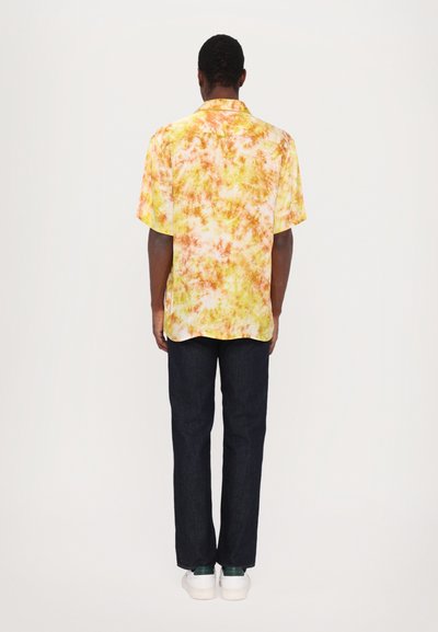 Camicetta a maniche corte in un vivace motivo tie-dye giallo, arancione e rosa, abbinata a pantaloni in denim scuro e sneakers bianchi.