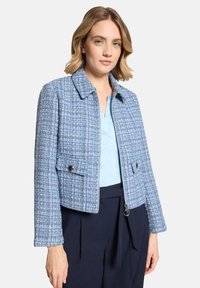 Femme portant une veste zippée texturée bleue par-dessus un haut bleu clair et un pantalon bleu marine foncé, debout contre un fond uni.