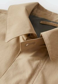 Camicia di cotone beige con colletto ampio, dotata di un bottone. Il tessuto è liscio con cuciture visibili lungo i bordi.