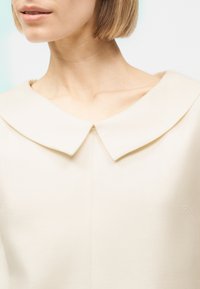 Γυναίκα με κοντά καστανά μαλλιά που φοράει μπλούζα κρεμ χρώματος με ευρύ, μυτερό γιακά, μπροστά σε φωτεινό φόντο.