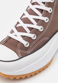 Brązowy tenisówka z płótna z białymi sznurowadłami, zaokrąglonym czubkiem, gumową podeszwą i teksturowanym, antypoślizgowym spodem. Srebrne przelotki i białe przeszycia jako detale.