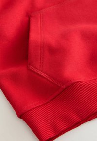 Roter Sweatshirt-Stoff mit weicher Textur, ausgestattet mit einer Seitentasche und gerippten Bündchen. Nahtdetails verbessern die gesamte Konstruktion.
