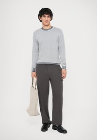 Αμάνικο πουλόβερ με ρίγες σε ναυτικό και λευκό χρώμα, συνδυασμένο με σκούρες γκρι παντελόνες και μαύρα παπούτσια. Το μοντέλο κρατά μια ανοιχτού μπεζ τσάντα.