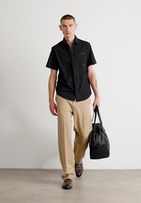 Cămașă neagră cu mâneci scurte, pantalonii bej cu picior larg, pantofi maro, purtând o geantă neagră tip tote. Design minimalist, culori uni.