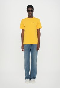 Tricou din bumbac galben cu guler rotund și un mic logo negru, asociat cu blugi decoltați largi de culoare albastră deschisă și adidași albi. Ochelari de soare negri purtați.