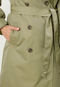 Oliwkowy płaszcz trench z podwójnymi guzikami, paskiem w talii i kieszeniami po bokach. Materiał gładki i lekko błyszczący.