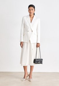 Kvinna med en vit texturerad kavaj och kjol, som håller en svart kviltad handväska med kedjearmar, stående mot en neutral bakgrund.