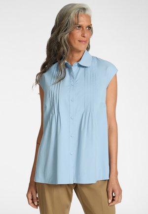 Kvinde med langt gråt hår iført en lyseblå ærmeløs skjorte med knapper og beige bukser, stående mod en hvid baggrund.