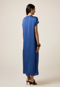 Donna in un abito blu reale lungo fino alla caviglia, sandali neri con tacco, e diversi braccialetti neri, in piedi di lato contro uno sfondo beige.