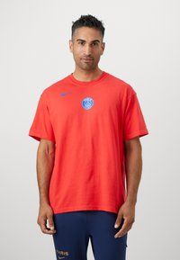 Rød bomulds T-shirt med korte ærmer, der har et blåt Nike-logo på venstre skulder og et cirkulært Paris-grafik i midten.