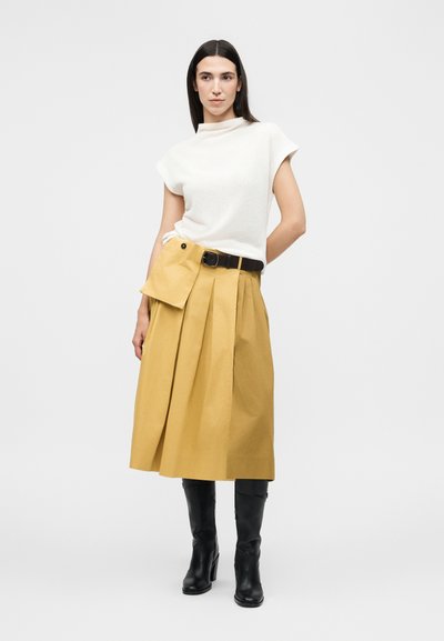 Γυναίκα με ίσια σκουρόχρωμα μαλλιά που φοράει λευκό μπλουζάκι χωρίς μανίκια, μουσταρδί πλισέ midi φούστα με προσαρτημένη πτέρυγα, μαύρη ζώνη και μαύρες μπότες μέχρι το γόνατο.