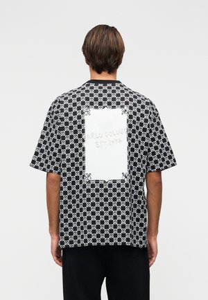 Mann trägt ein kurzärmeliges Hemd mit schwarz-weiß kariertem Muster, zirkulären Logos und einem großen weißen rechteckigen Aufnäher auf der Rückseite.