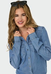 Chemise en denim bleu clair avec deux poches sur la poitrine et fermeture à boutons, dotée de manches retroussées et d'une coupe décontractée. Lunettes de soleil reposant sur la tête.