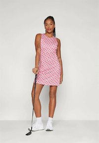 Sukienka golfowa w kolorze różowym z plecionym wzorem. Bez ramiączek, do kolan. Modelka trzyma kij golfowy, nosi białe trampki i skarpetki.