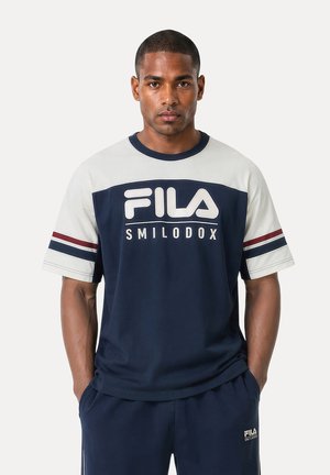 Mand iført navy og hvid FILA Smilodox t-shirt og navy bukser med hænderne i lommerne, vendt fremad mod en ensfarvet baggrund.