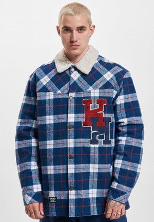 Blue and white plaid shirt jacket with a cream sherpa collar, featuring a red and navy embroidered logo on the chest and metal buttons.