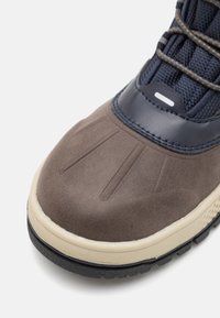 Acercamiento a la punta de una bota impermeable marrón y azul marino con una plantilla beige y una suela negra resistente sobre un fondo blanco.