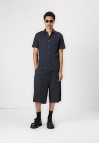Chemise à manches courtes, bleu marine foncé, avec un bouton et une poche ; associée à un short large gris foncé arrivant au genou ; bottes noires ; lunettes de soleil portées.