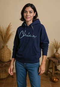 Navy blauwe hoodie met "Chica" in lichtblauwe cursieve letters, voorzien van een voorzak en een capuchon met koord, gecombineerd met blauwe jeans.