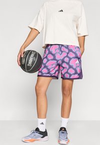 Μαύρο και ροζ γραφικό τύπωμα μπάσκετ σορτς με ελαστική μέση, σε συνδυασμό με μια μπεζ υπερμεγέθη μπλούζα και μαύρα αθλητικά παπούτσια.