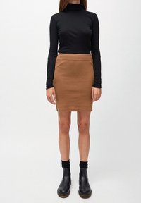 Vrouw in een zwarte longsleeve turtleneck, bruine rok tot de knie, zwarte sokken en zwarte enkellaarsjes, staande tegen een effen achtergrond.