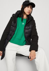 Veste noire et bouffante avec une capuche, portée sur un sweatshirt vert et un jean blanc. Présente un design matelassé et une capuche avec cordon de serrage.