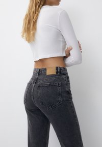 Haut blanc à manches longues cropped associé à un jean en denim noir taille haute, avec une étiquette en cuir marron sur la ceinture arrière et une finition texturée.