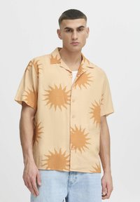 Kurzärmliges, leichtes Hemd aus beigem Stoff mit großen orangen Sonnenmustern. Verfügt über einen Kragen und eine Knopfleiste, kombiniert mit Jeans.